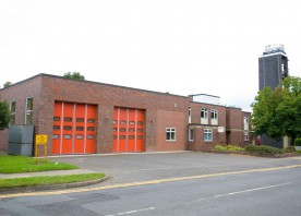 Elm Lane fire station. This is a brick built building, surrounded by grass and trees. You can see two big red garage doors, and a firefighter training tower in the background.
