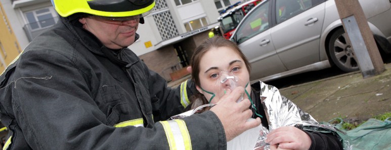 A firefighter wearing black fire kit and a yellow helmet holds an oxygen mask to a lady's face. In the background is a block of flats.