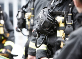 A breathing apparatus mask hangs down from a firefighters neck. You can see the mask, up close, and can just make out a number of firefighters gathered round the individual's mask. They are wearing black firefighting kit.