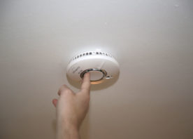 A smoke alarm is fitted to a ceiling. A finger is pushing the test button in the middle.