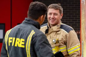 Two firefighters talk in front of a red door. One is wearing light brown kit, and is smiling towards the camera. The other is looking away, and is wearing dark grey fire kit.