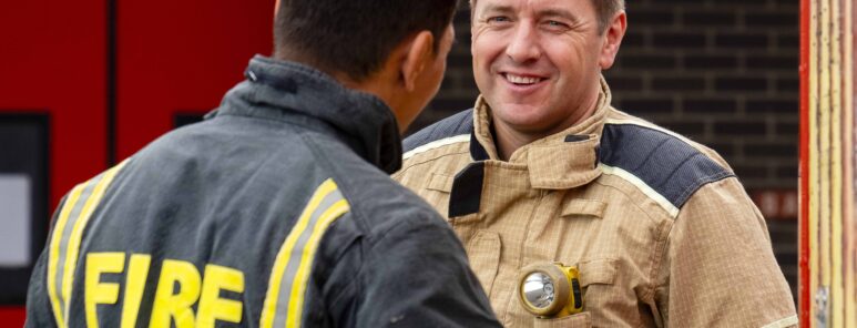 Two firefighters talk in front of a red door. One is wearing light brown kit, and is smiling towards the camera. The other is looking away, and is wearing dark grey fire kit.