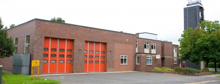 Elm Lane fire station. This is a brick built building, surrounded by grass and trees. You can see two big red garage doors, and a firefighter training tower in the background.