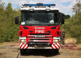 The front of a big red fire engine, in the middle of a forest. You can see two firefighters talking towards the rear of the appliance.