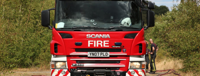 The front of a big red fire engine, in the middle of a forest. You can see two firefighters talking towards the rear of the appliance.