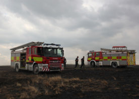 Two fire engines are parked up on an area of farm land, on a hill. The weather is dull and it appears to be in the late evening. Two firefighters can be seen in the distance, walking between the two fire engines.
