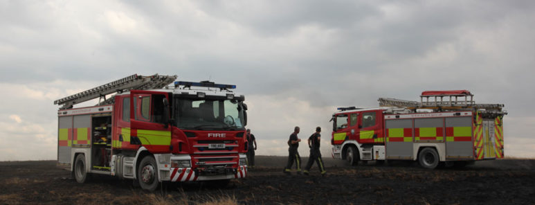 Two fire engines are parked up on an area of farm land, on a hill. The weather is dull and it appears to be in the late evening. Two firefighters can be seen in the distance, walking between the two fire engines.