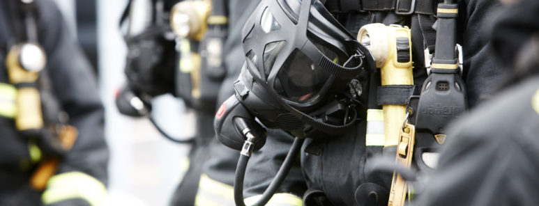 A breathing apparatus mask hangs down from a firefighters neck. You can see the mask, up close, and can just make out a number of firefighters gathered round the individual's mask. They are wearing black firefighting kit.