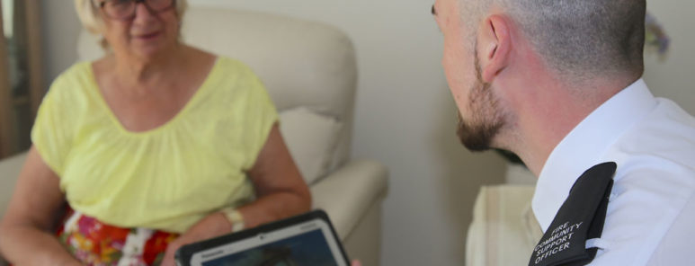 A fire safety officer holds a computer tablet as he sits and speaks to a female resident. She is wearing a yellow shirt, whilst he is wearing a white shirt.