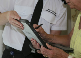 A fire safety officer, in a white shirt and tie, shows something to a member of the public on a computer tablet. You can't see their faces, but the lady he is speaking to is wearing yellow.