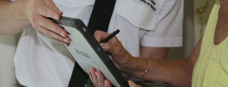 A fire safety officer, in a white shirt and tie, shows something to a member of the public on a computer tablet. You can't see their faces, but the lady he is speaking to is wearing yellow.