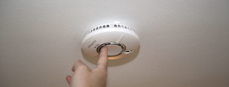 A smoke alarm is fitted to a ceiling. A finger is pushing the test button in the middle.