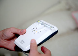 Two hands hold a carbon monoxide alarm. The alarm is white with a series of small LED lights on the front.