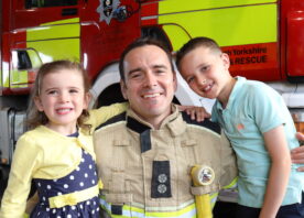 A firefighter poses with his son and daughter as part of a campaign photo shoot. He is wearing fire kit. All three are smiling, in front of a fire engine.
