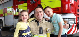 A firefighter poses with his son and daughter as part of a campaign photo shoot. He is wearing fire kit. All three are smiling, in front of a fire engine.