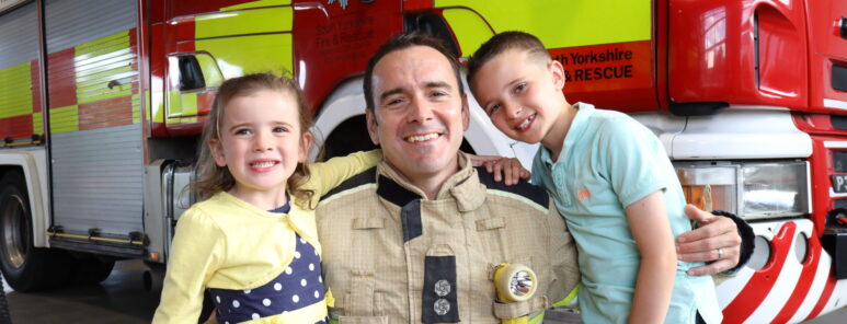 A firefighter poses with his son and daughter as part of a campaign photo shoot. He is wearing fire kit. All three are smiling, in front of a fire engine.