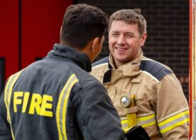 Two firefighters talk in front of a red door. One is wearing light brown kit, and is smiling towards the camera. The other is looking away, and is wearing dark grey fire kit.