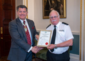 Two men pose together holding an award. One is wearing a black suit with a red tie, the other is wearing a white fire service shirt with a black tie.