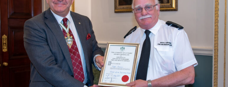 Two men pose together holding an award. One is wearing a black suit with a red tie, the other is wearing a white fire service shirt with a black tie.