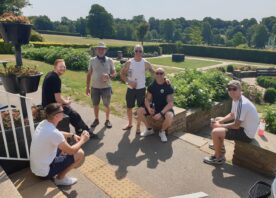 A group of firefighters, dressed mostly in shorts and t-shirts, gather for a photo. They are smiling and the sun is shining, with a background of green grass and trees.