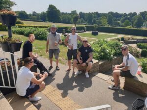 A group of firefighters, dressed mostly in shorts and t-shirts, gather for a photo. They are smiling and the sun is shining, with a background of green grass and trees.