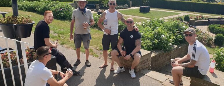 A group of firefighters, dressed mostly in shorts and t-shirts, gather for a photo. They are smiling and the sun is shining, with a background of green grass and trees.